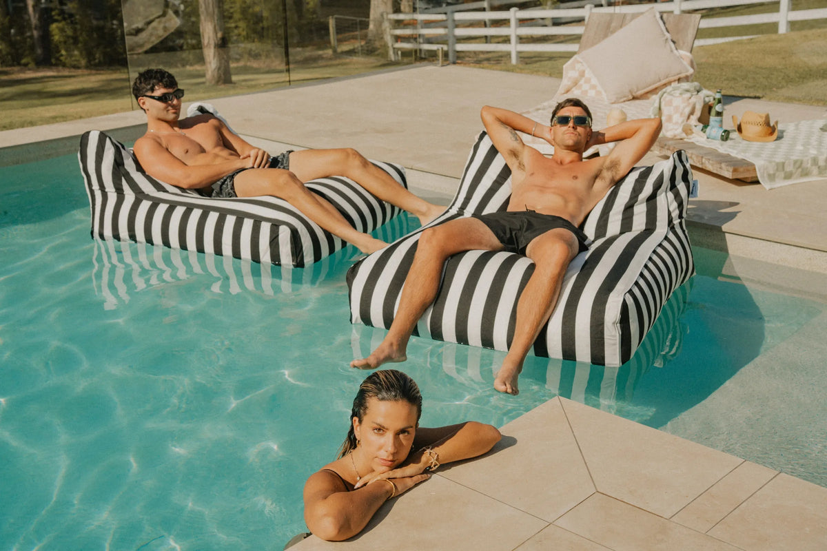 Two men relaxing on black and white striped floating loungers in a luxury pool under the sun, with a woman resting on the pool edge  House of Dune Orca Stripe Floating Lounger for poolside leisure.