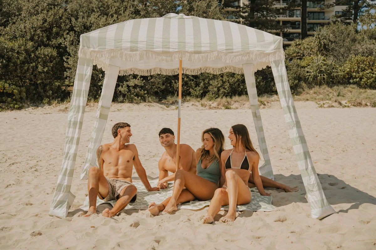 Group of friends sitting under a pastel green and white striped House of Dune Salted Sage Cabana on a sunny beach, enjoying shade and coastal views in a premium beach canopy setup.