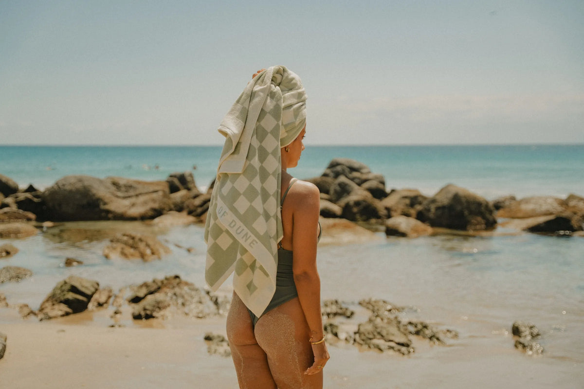 Beach model standing near ocean rocks wearing a green swimsuit and wrapped in a checkered pastel green House of Dune Salted Sage Towel, highlighting a luxury coastal lifestyle scene.