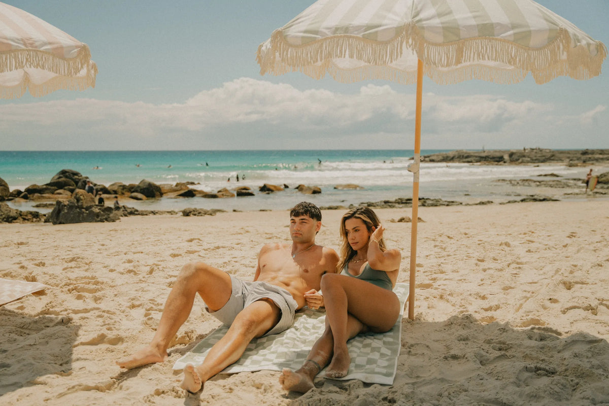 Couple relaxing under a mint-green and white striped House of Dune Salted Sage Beach Umbrella by the ocean while sitting on a checkered towel, providing premium coastal sun protection.