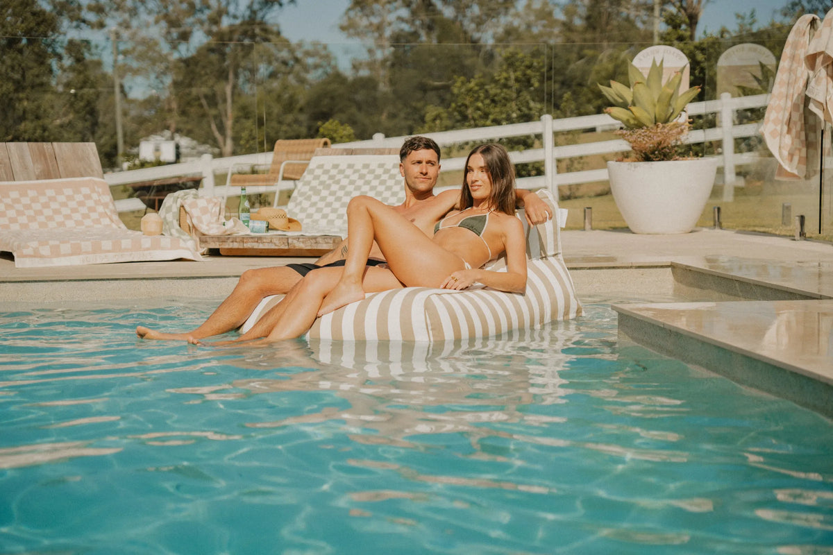Couple relaxing on a beige and white striped House of Dune Sand Drift Floating Lounger in a luxury poolside setting, highlighting a modern resort lifestyle with a floating pool chair.