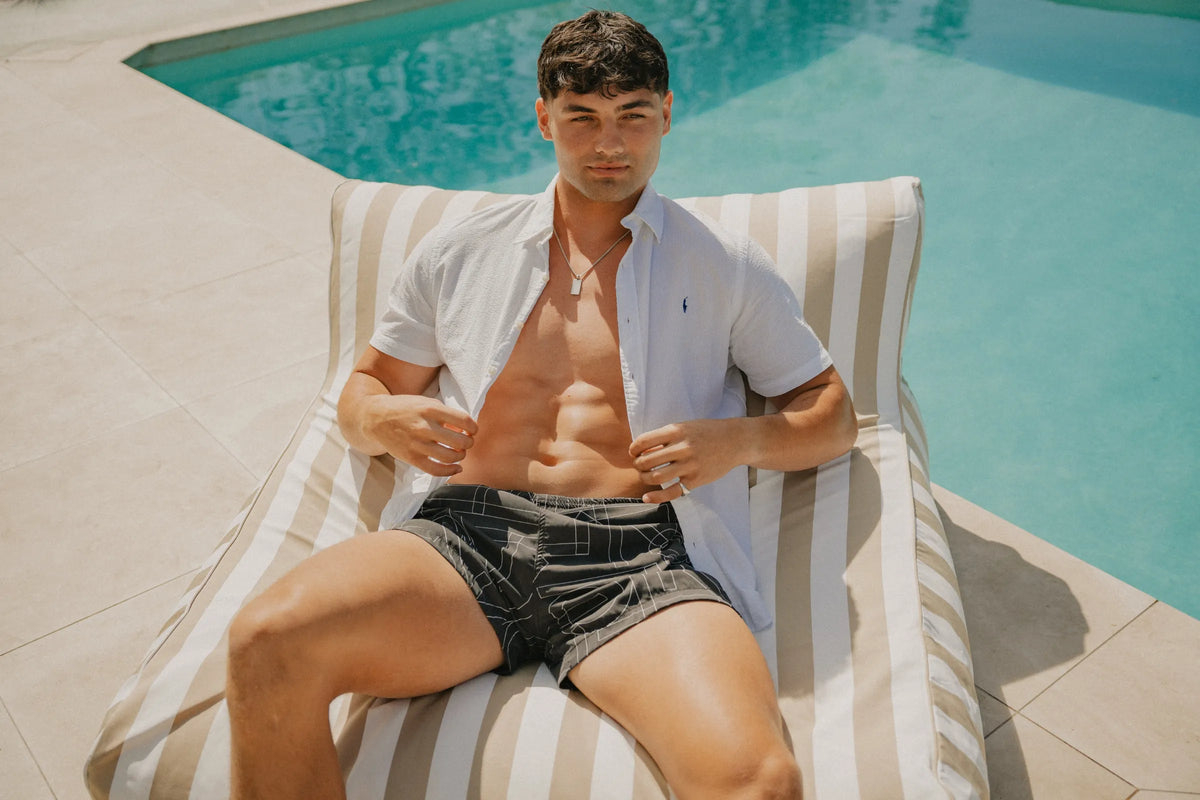 Man lounging on a beige and white striped House of Dune Sand Drift Floating Lounger beside a pool under the sun, featuring stylish summer poolside furniture.