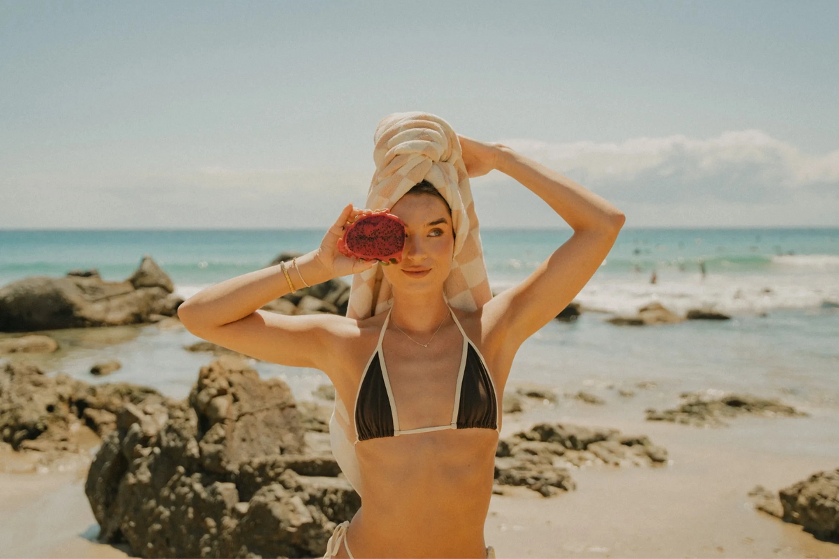 Model standing on the beach with a checkered beige House of Dune Sand Drift Towel wrapped around her head while holding a dragon fruit, creating a relaxed coastal lifestyle look.
