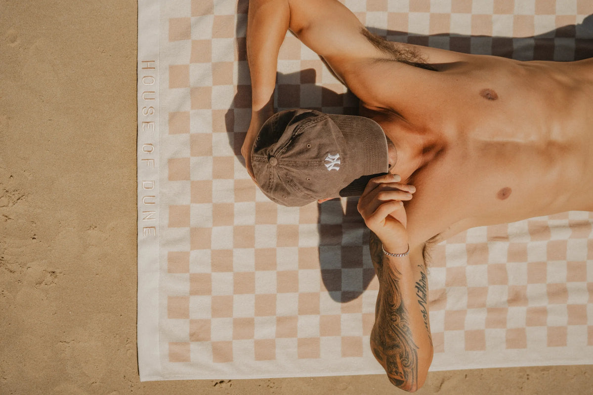 Man sunbathing on sand using a checkered beige House of Dune Sand Drift Towel with embroidered logo, showcasing a luxury cotton beach towel design.