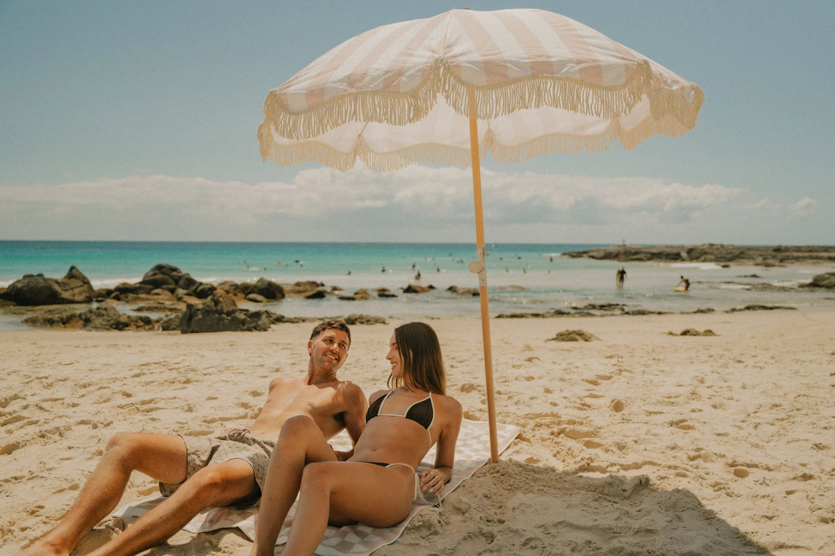 Couple relaxing on the sand under a cream and beige striped House of Dune beach umbrella with fringe detail, surrounded by rocks and turquoise water on a sunny coastal day.