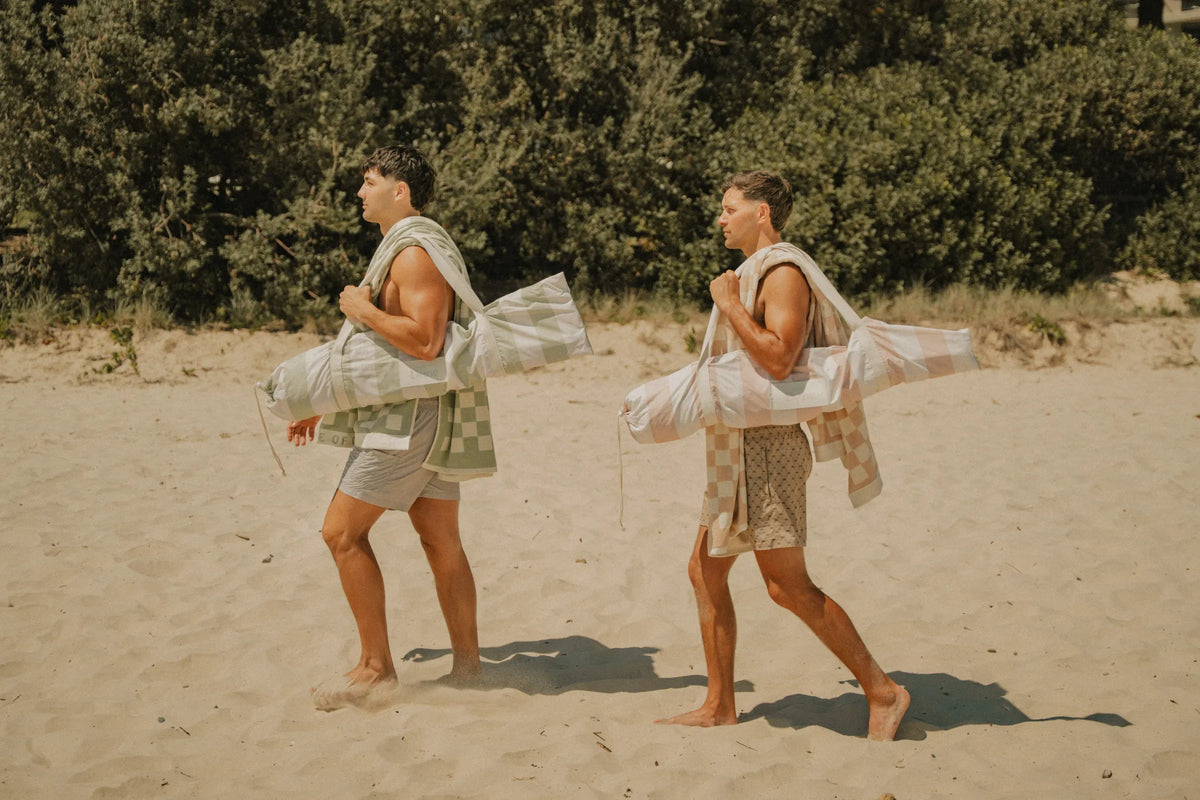 Two men carrying House of Dune Salted Sage Beach Umbrellas and towels along the sand, demonstrating lightweight and portable summer beach gear with a relaxed coastal aesthetic.