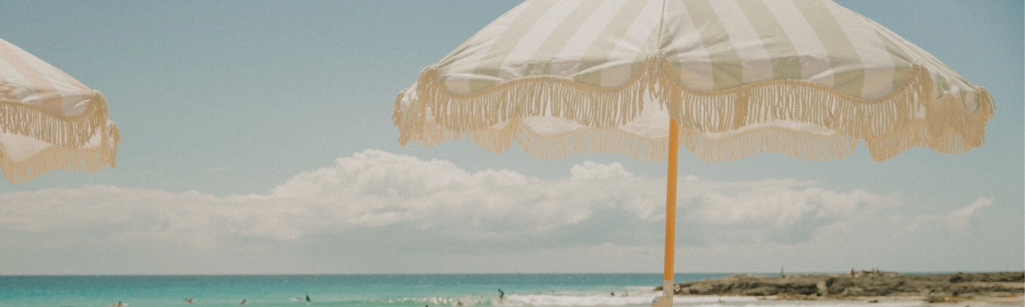 Top-down view of four House of Dune striped beach umbrellas in alternating beige and green with cream fringe, shading sunbathers on sandy beach with towels and surfboard.