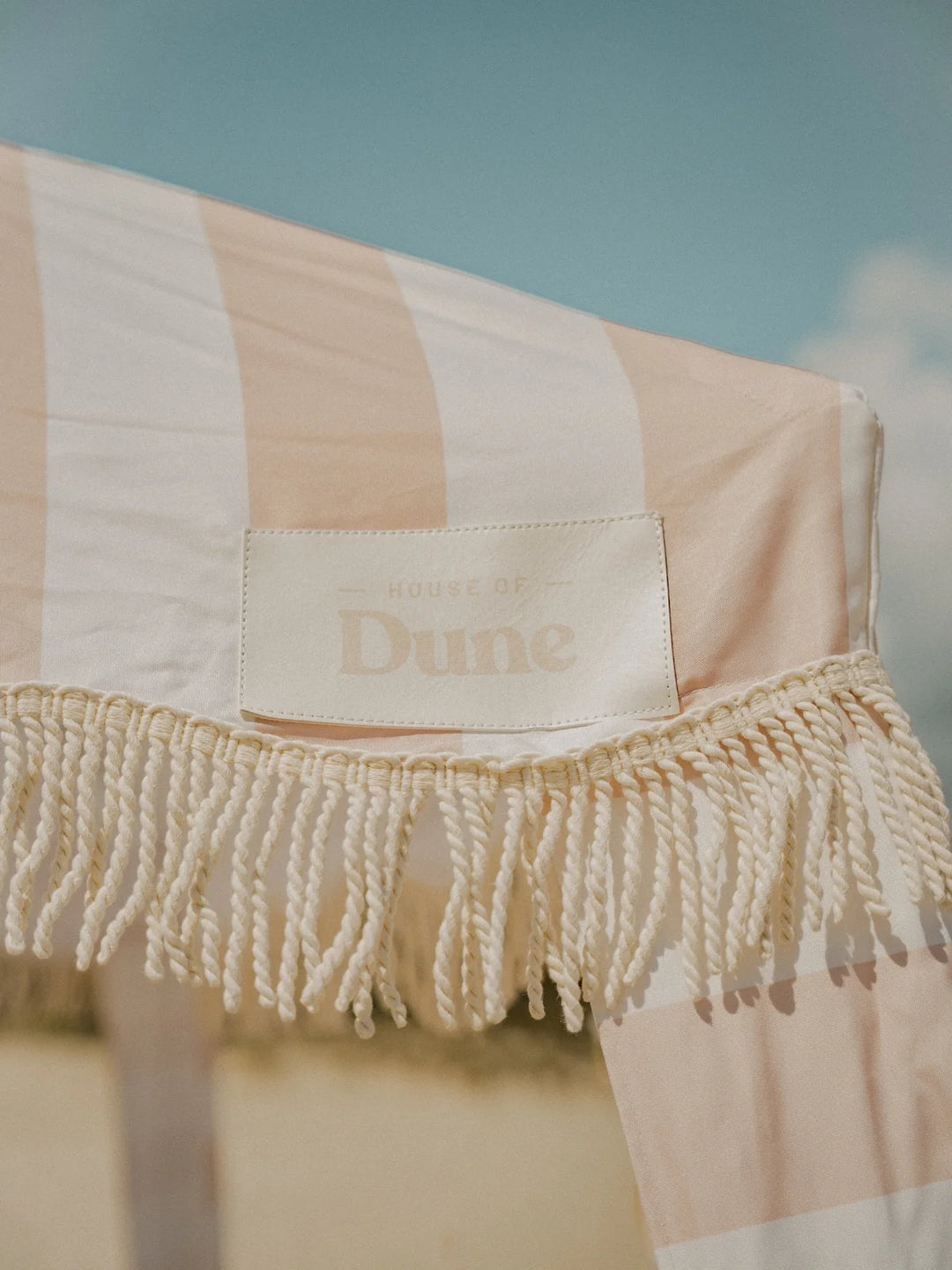 Close-up of the House of Dune Sand Drift Cabana canopy showing beige and white stripes, soft cotton fringe trim, and the stitched House of Dune logo against a blue sky.