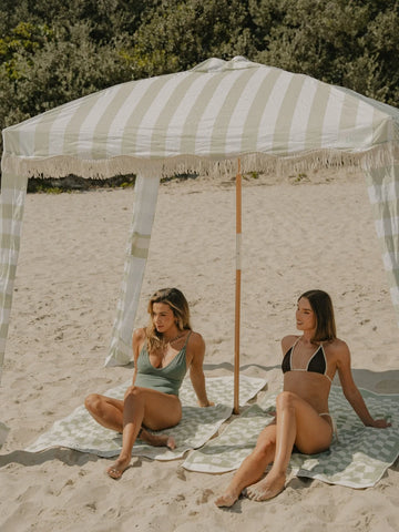 Two women relax beneath the House of Dune Salted Sage Cabana on a sandy beach, lounging on matching checkered towels. The scene captures a calm coastal moment under the soft green and white striped shade