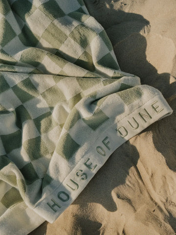 Close-up of the Salted Sage checkered beach towel with embossed House of Dune logo resting on warm sand.