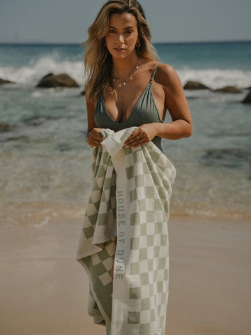 Woman in green swimsuit standing on the beach holding a Salted Sage checkered towel by House of Dune with waves behind her.