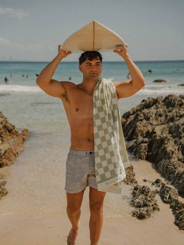 Man carrying a surfboard on the beach with a Salted Sage checkered towel draped over his shoulder, House of Dune.