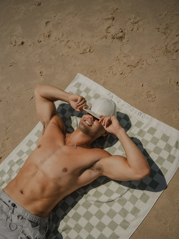 Man relaxing on the sand wearing grey shorts, lying on the Salted Sage checkered towel by House of Dune.