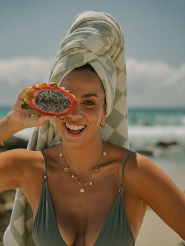 Smiling woman holding a slice of dragon fruit while wearing a Salted Sage checkered towel wrapped around her head, House of Dune.
