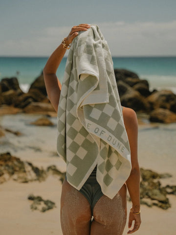 Back view of woman drying her hair with a Salted Sage checkered towel near ocean rocks, House of Dune coastal scene.