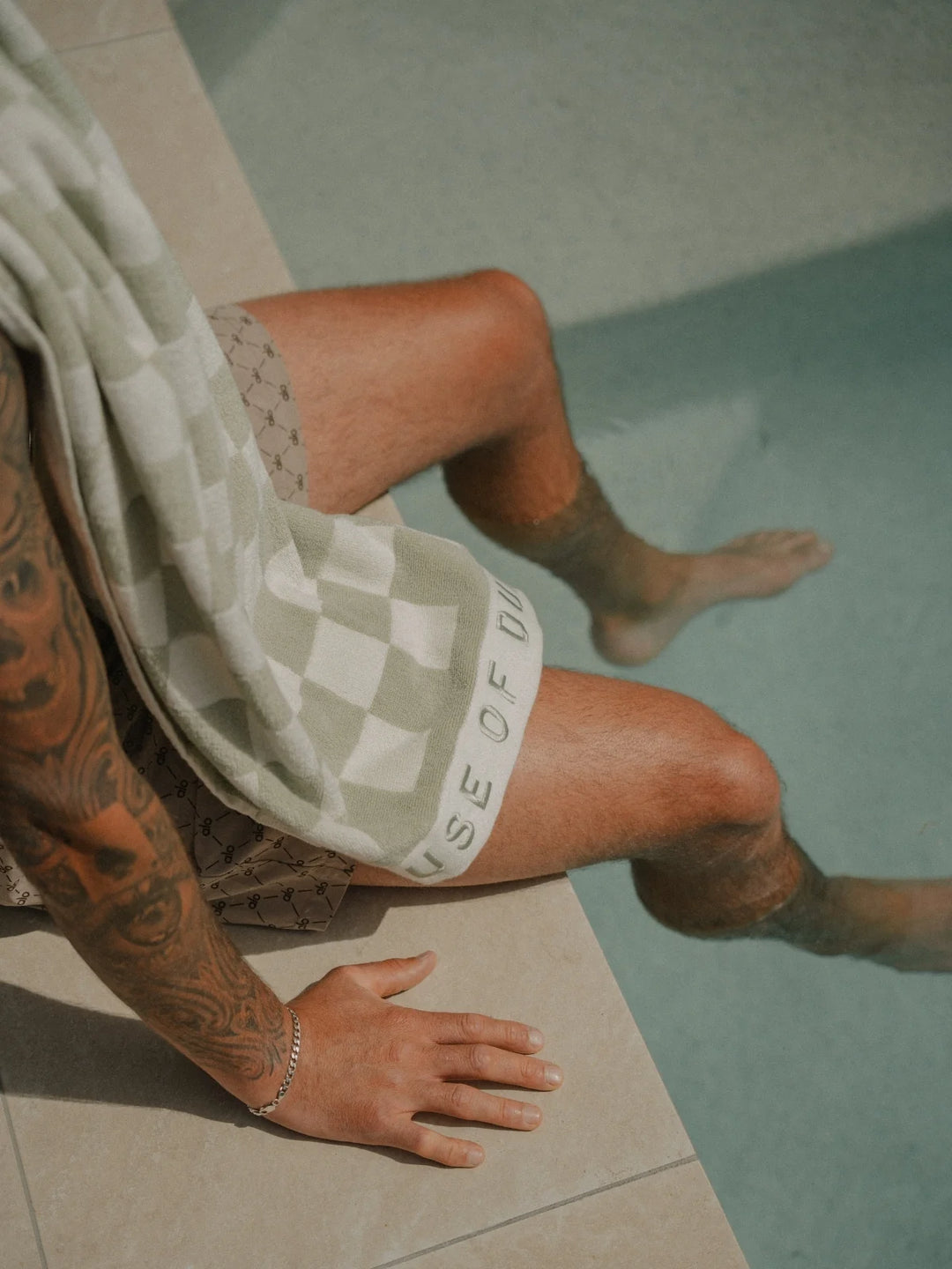 Man sitting beside a swimming pool with a Salted Sage checkered towel draped across his lap, House of Dune summer collection.