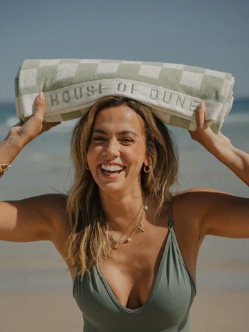Smiling woman in green swimsuit holding a rolled Salted Sage checkered beach towel by House of Dune.