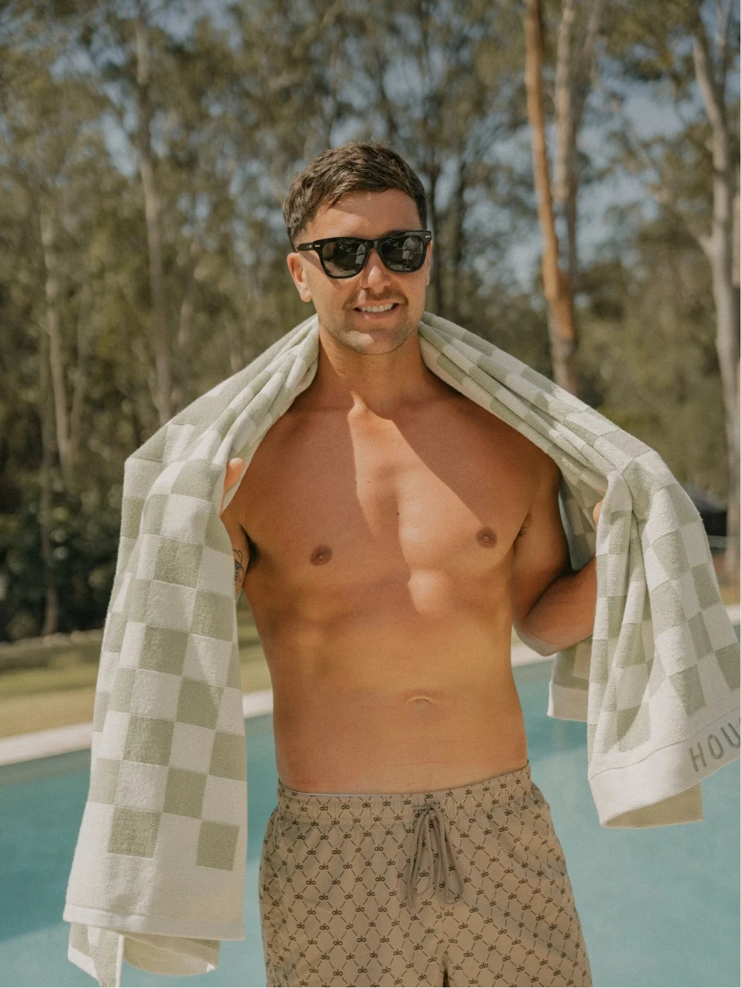 Man standing by the pool wearing sunglasses and draped in a Salted Sage checkered towel from House of Dune.