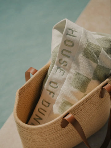 Folded Salted Sage checkered towel with House of Dune logo tucked inside a woven straw beach bag.