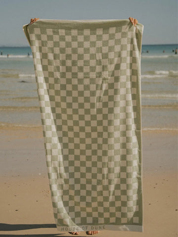 Salted Sage checkered beach towel held upright on sandy shore with the ocean in the background.