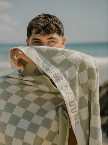 Man holding a Salted Sage checkered towel by House of Dune against a sunny coastal backdrop.