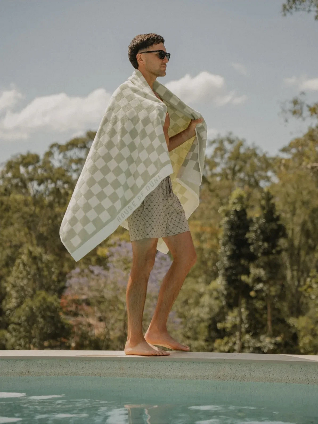 Man with sunglasses draped in the Salted Sage checkered towel walking beside a pool, House of Dune collection.
