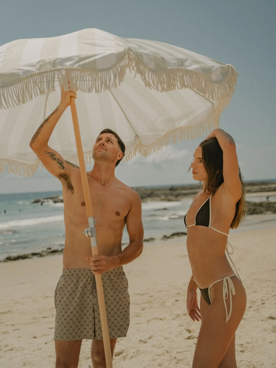 Man and woman assembling the Salted Sage striped beach umbrella from House of Dune on a golden sandy beach.