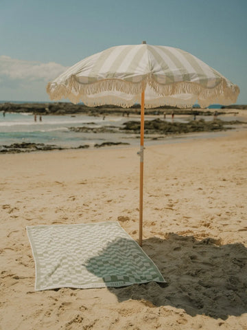 Salted Sage beach umbrella providing shade over a checkered House of Dune towel at the shoreline.