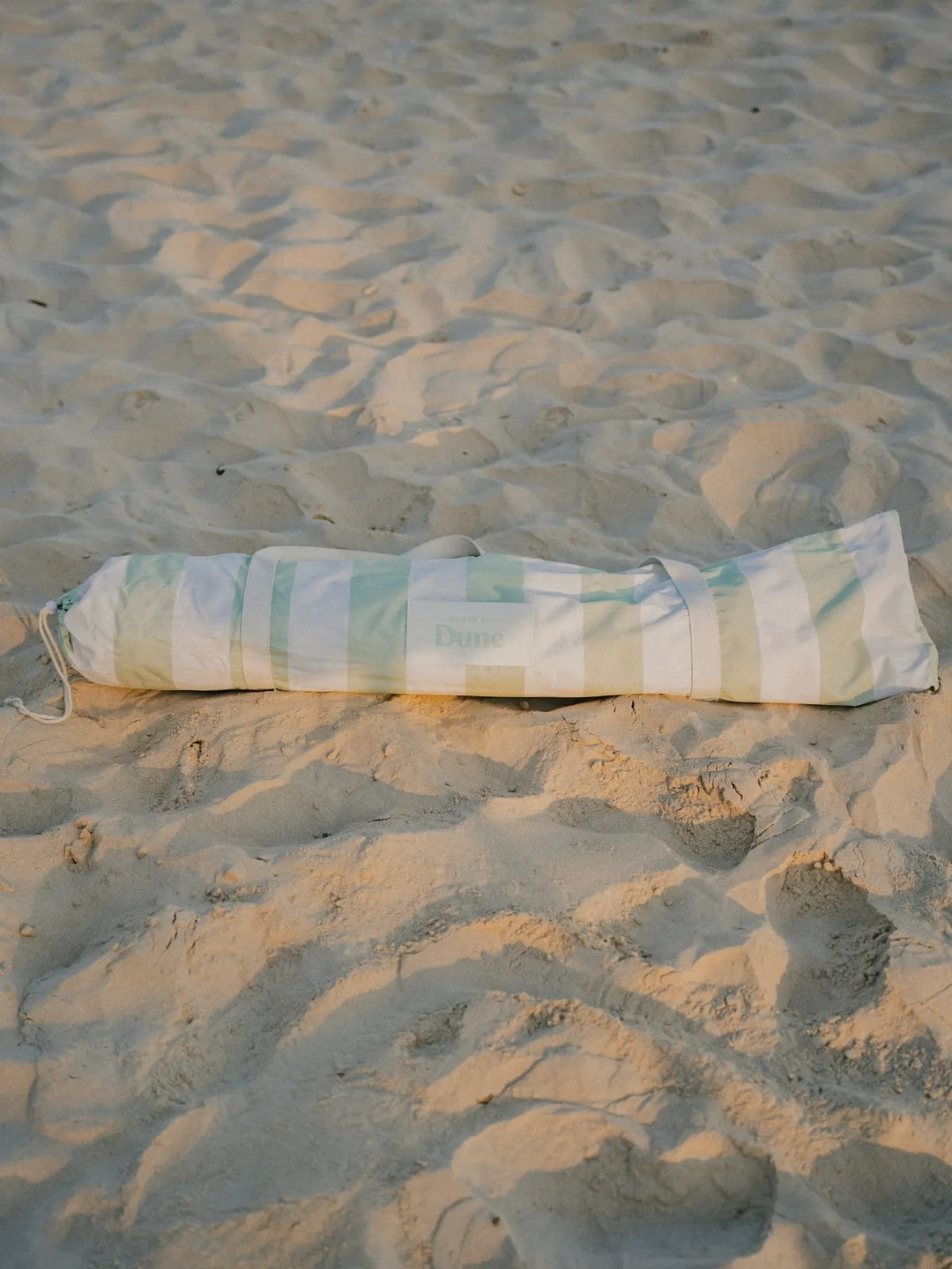 Salted Sage beach umbrella neatly packed inside its striped House of Dune carry bag resting on sand