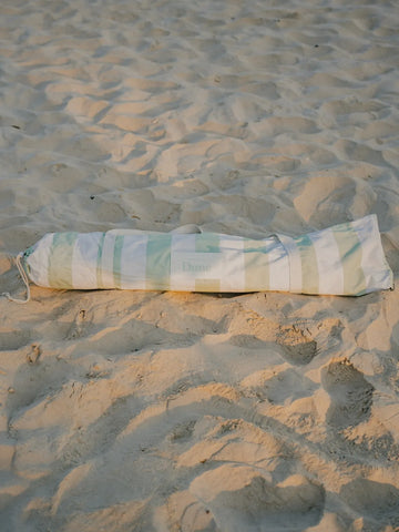 Salted Sage beach umbrella neatly packed inside its striped House of Dune carry bag resting on sand