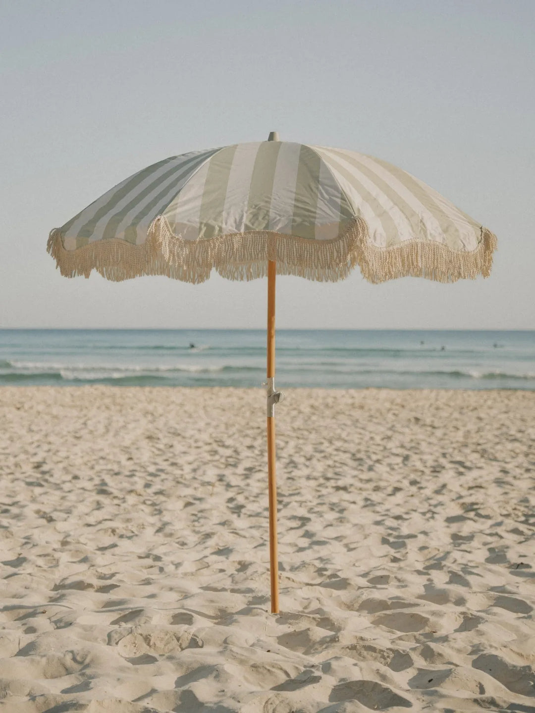 Salted Sage beach umbrella with cream fringe open on the beach under soft morning light by House of Dune.