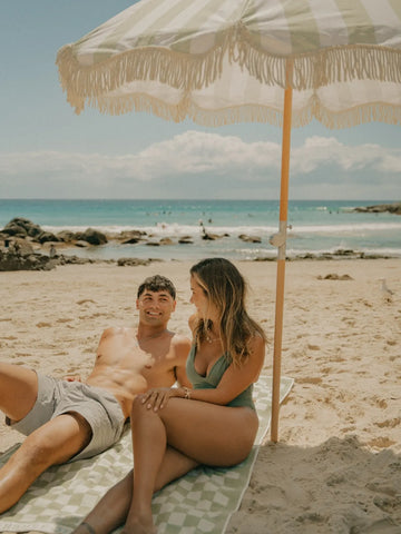 Couple relaxing together under the open Salted Sage beach umbrella by House of Dune with ocean waves behind them.