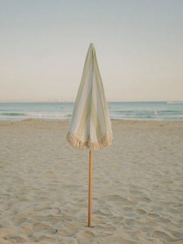Salted Sage striped beach umbrella folded upright in the sand with calm ocean backdrop, by House of Dune.
