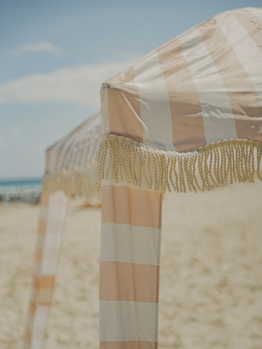 Detail shot of the House of Dune Sand Drift Cabana with wide neutral stripes and knotted fringe, capturing the soft coastal tones of the beach setting.