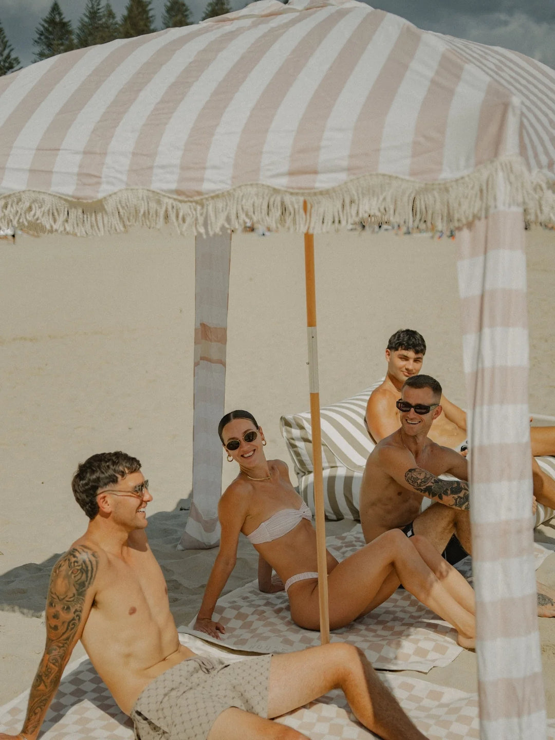 Group of friends lounging beneath the House of Dune Sand Drift Cabana, surrounded by sand and sunshine, highlighting its classic striped design and airy shade.