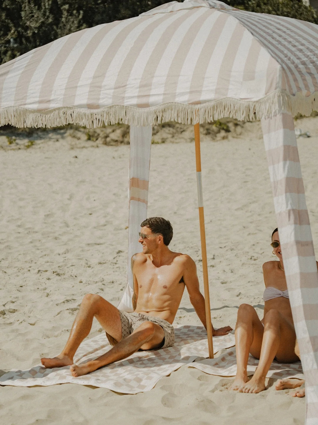 Couple relaxing under the House of Dune Sand Drift Cabana on the beach, sitting on striped towels beneath the soft beige and white canopy.