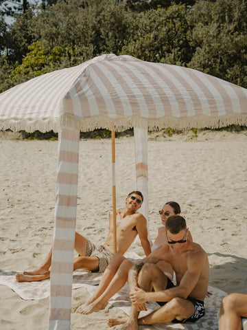 Friends gathered beneath the House of Dune Sand Drift Cabana, enjoying a relaxed beach afternoon with coastal tones and striped fabric providing elegant shade.