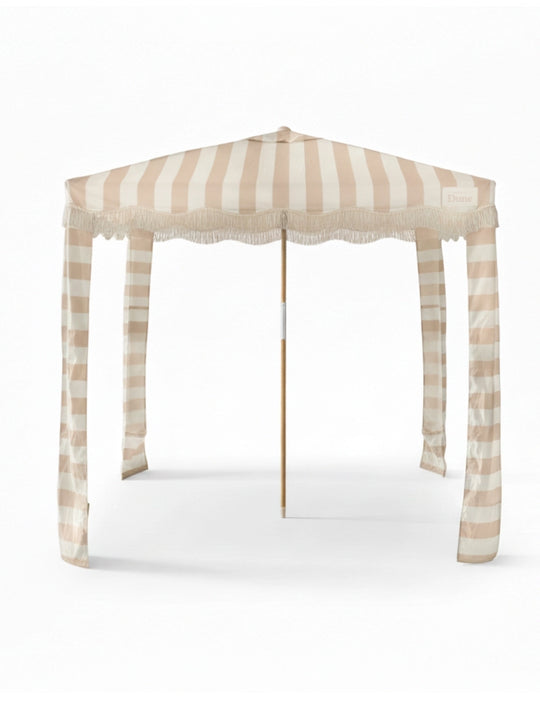Front view of the House of Dune Sand Drift Cabana, showcasing its beige and white striped canopy with fringe trim, wooden pole, and draped corner panels designed for modern coastal shade.