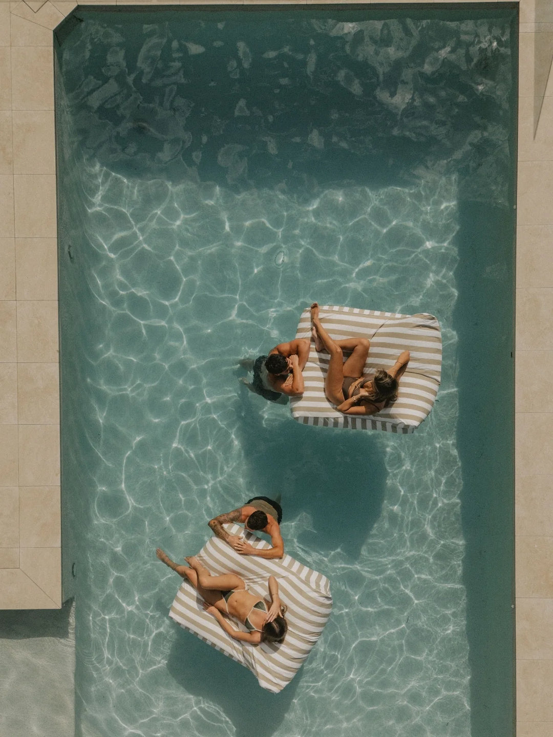 Aerial view of three House of Dune Sand Drift Floating Loungers in a sunlit pool with friends relaxing, showcasing the elegant beige and white striped design.
