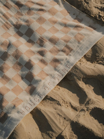 Close-up of Sand Drift checkered beach towel with embossed House of Dune logo resting on soft golden sand.