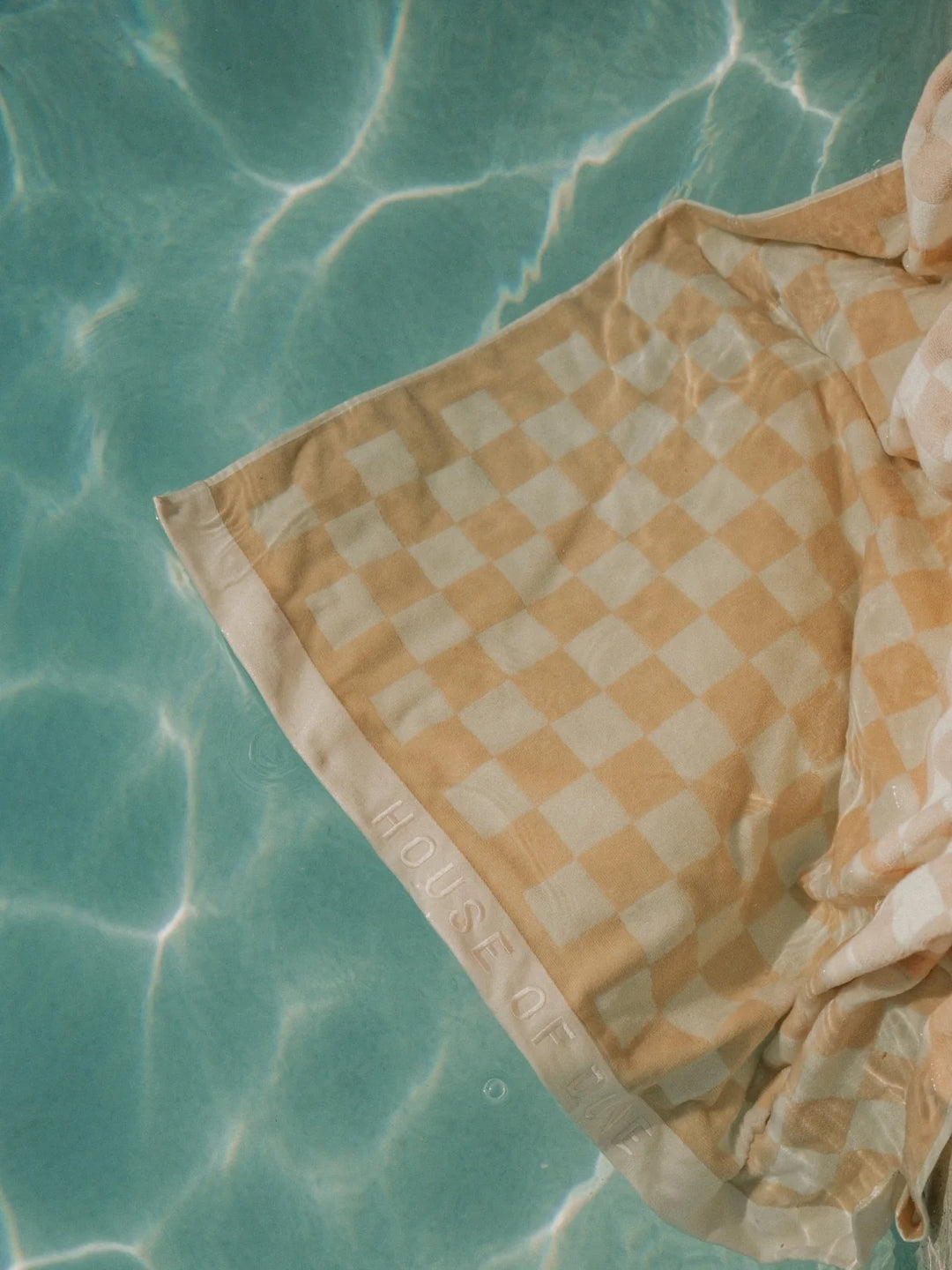 Sand Drift checkered towel by House of Dune partially submerged in clear turquoise pool water under sunlight.