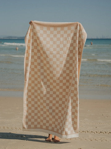 Sand Drift checkered beach towel held upright on the shoreline with the ocean waves in the background, House of Dune.