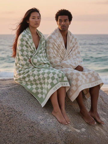 Two people wrapped in House of Dune checkered towels sitting on a rock at the beach during sunset, coastal tones.