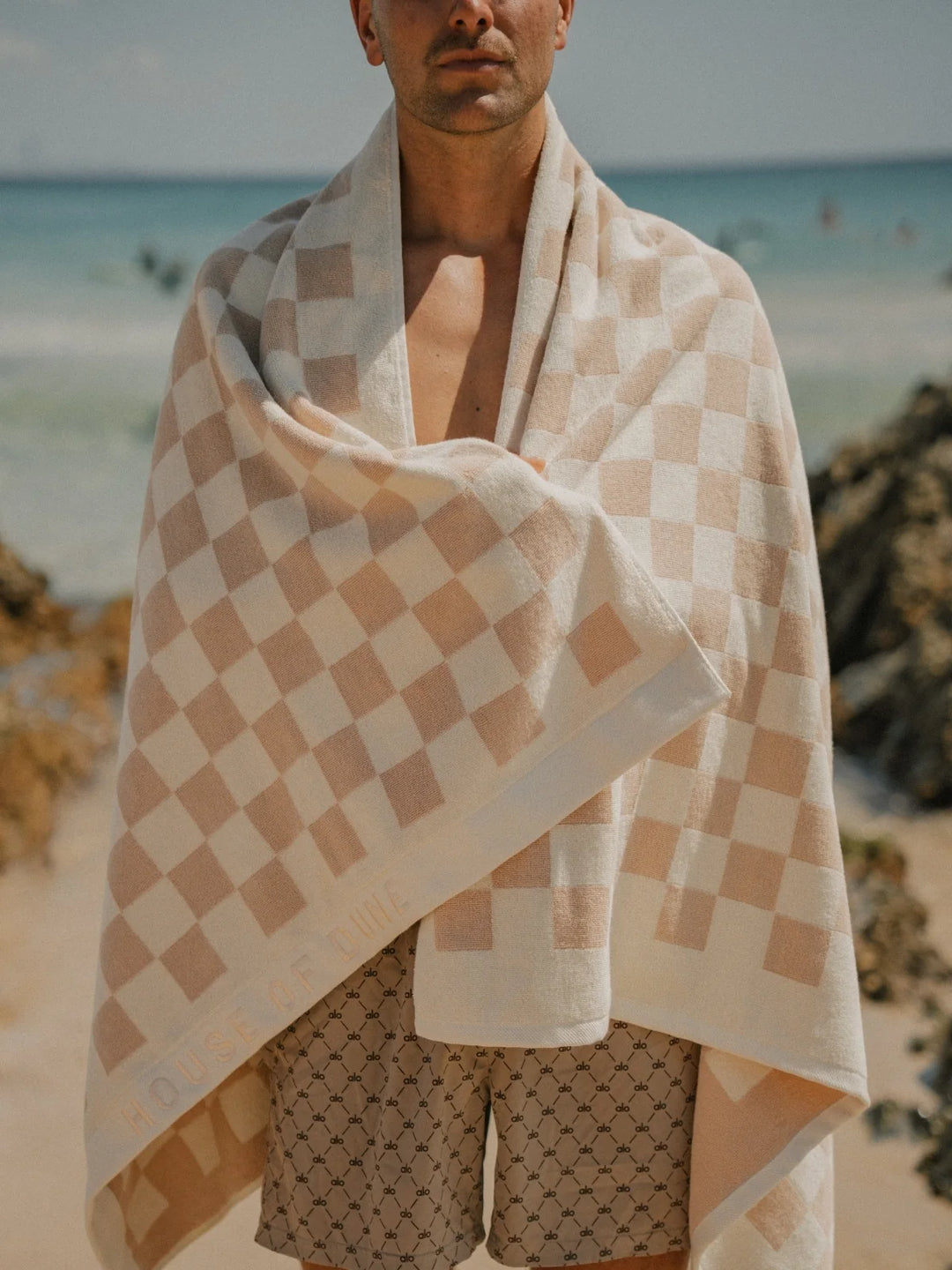Man standing on the beach with Sand Drift checkered towel draped over shoulders, House of Dune coastal lifestyle.