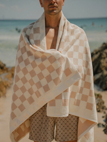 Man standing on the beach with Sand Drift checkered towel draped over shoulders, House of Dune coastal lifestyle.