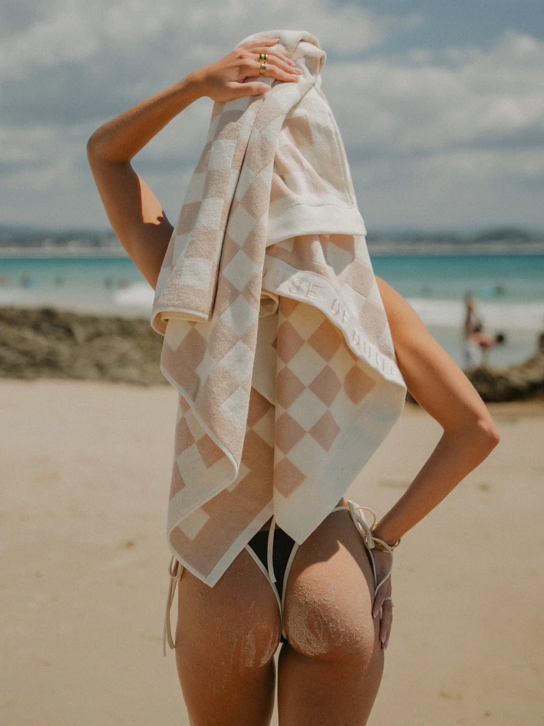 Back view of woman drying her hair with a Sand Drift checkered towel by the ocean, House of Dune coastal collection.