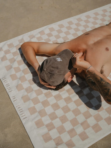Man lying on sand wearing cap and shorts, sunbathing on a Sand Drift checkered towel by House of Dune.