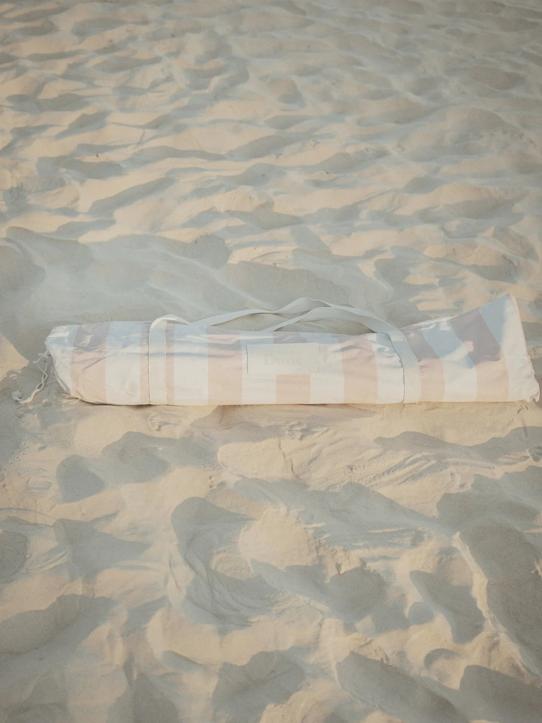 Sand Drift beach umbrella neatly packed in its striped House of Dune carry bag resting on soft sand.