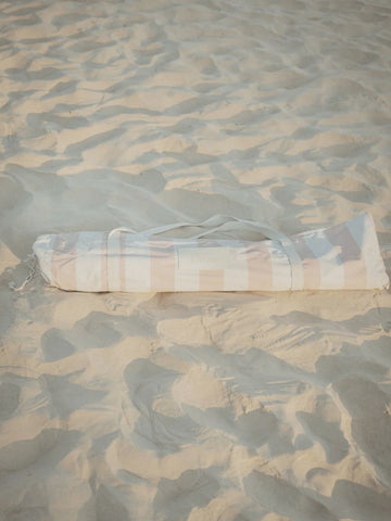 Sand Drift beach umbrella neatly packed in its striped House of Dune carry bag resting on soft sand.