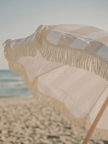 Sand Drift beach umbrella with soft beige stripes open on the beach in warm afternoon light, House of Dune.